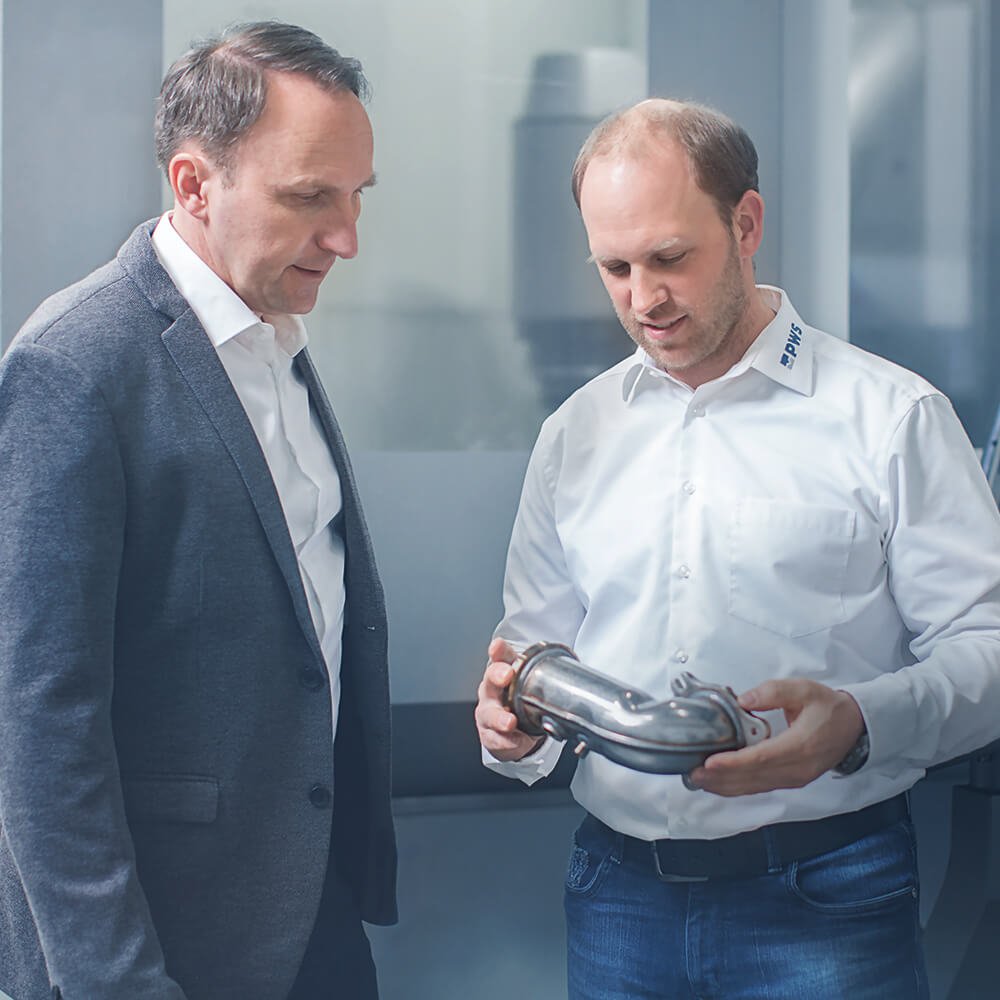 Zwei Männer, die ein Werkstück aus Metall betrachten, das einer von ihnen in den Händen hält.