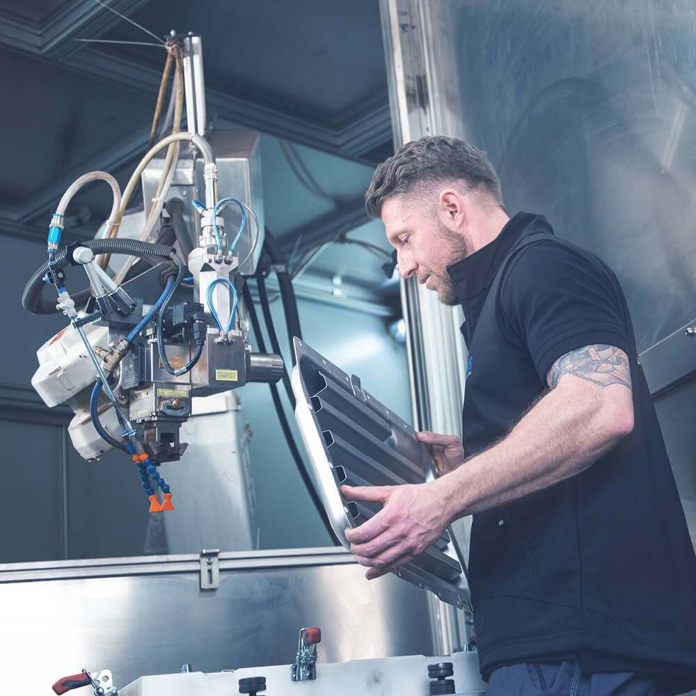 Mann hält ein Werkstück aus Metall vor einer Maschine.